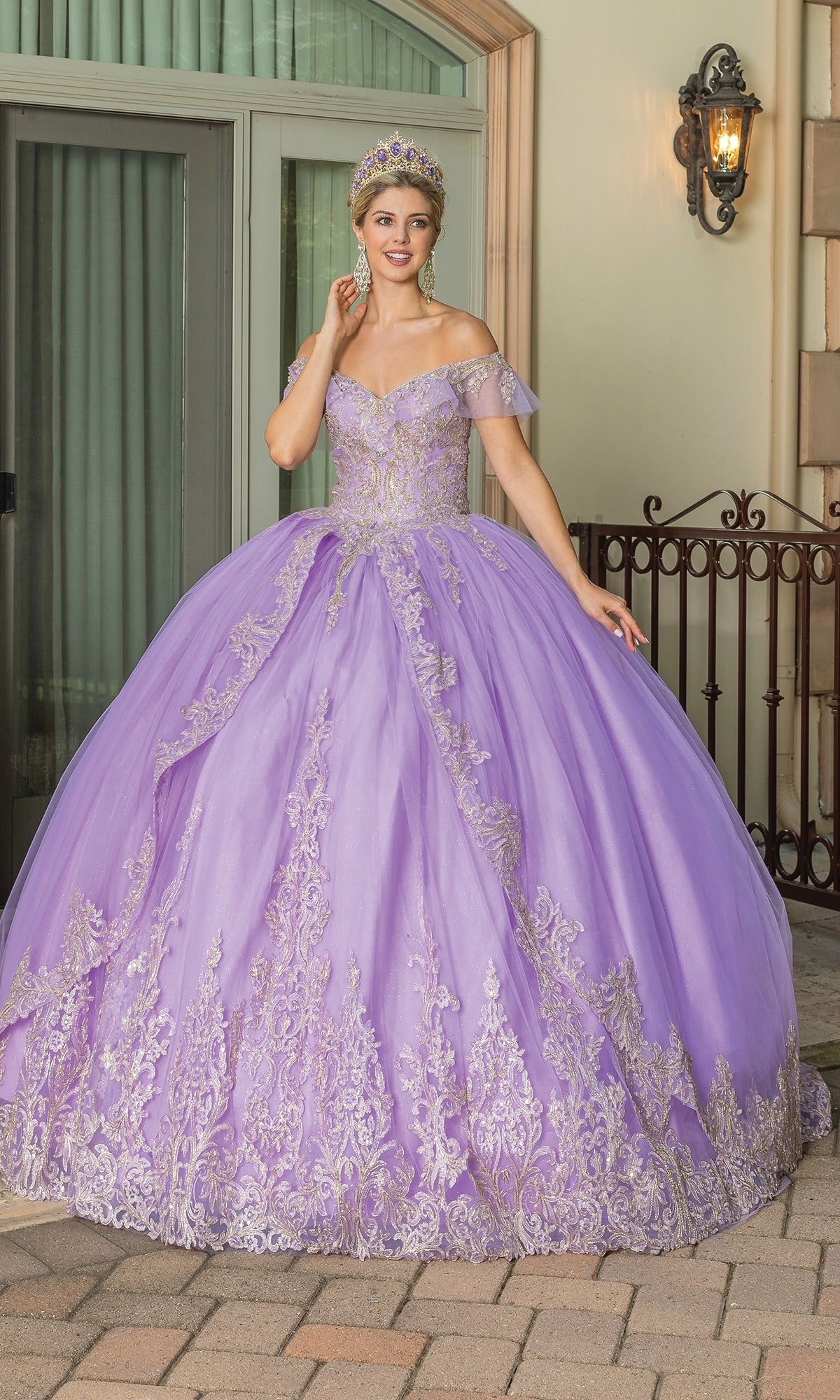 A woman stands outside in the Off-the-Shoulder Quinceanera Dress 1615, a lavender a-line ball gown with gold lace embroidery, an off-the-shoulder neckline, and a matching jeweled tiara.